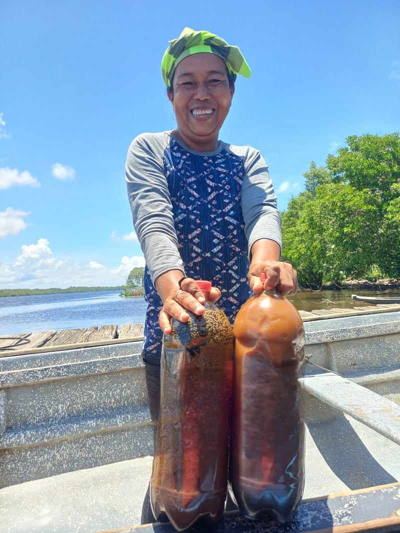 Smiling Miskito woman on a boat holding two bottles of batana oil with river and mangroves behind her